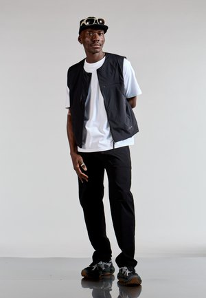 Chaleco ligero negro sobre una camiseta blanca, combinado con pantalones negros y zapatillas grises con un patrón de camuflaje. Los accesorios incluyen una gorra negra con gafas de sol.