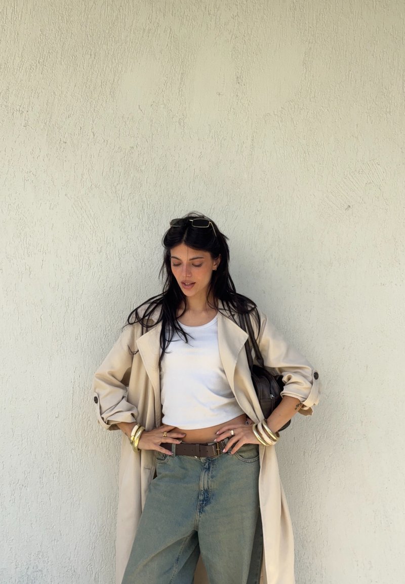 Woman with long dark hair wearing a beige trench coat, white crop top, blue jeans, gold bracelets, and sunglasses on her head poses against a textured wall.