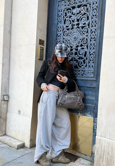 Mujer con pantalones grises holgados, chaqueta negra y gorra de cuadros mirando su teléfono, de pie junto a una puerta azul ornamentada con un panel de latón y el número 32.