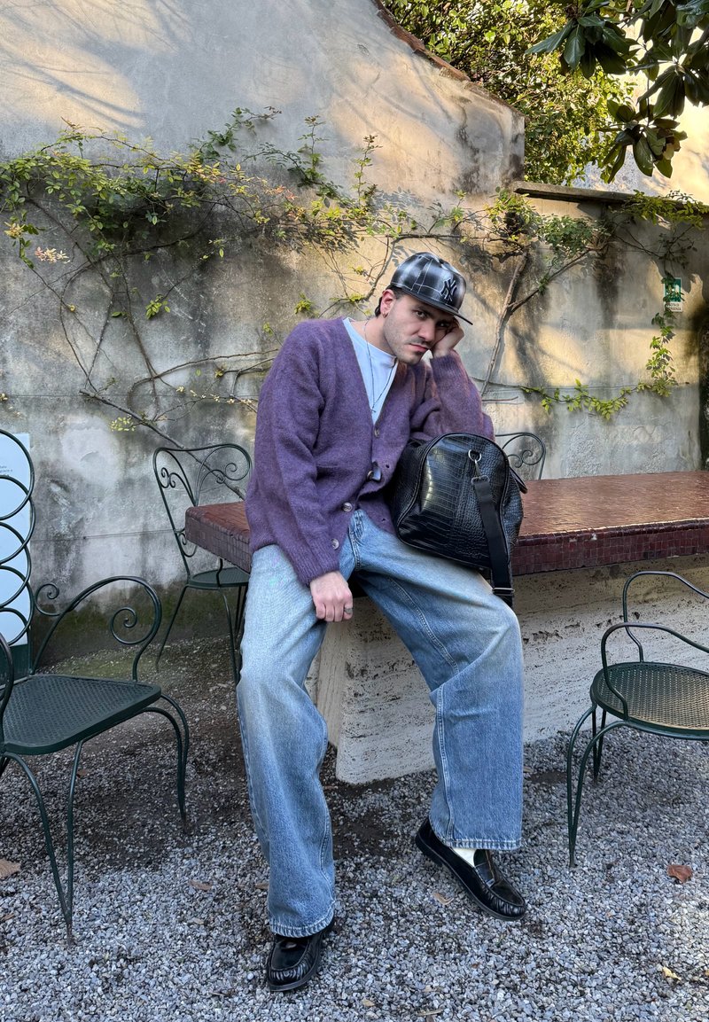 Jeune homme en cardigan violet et casquette assis sur un banc extérieur avec un sac noir, entouré de chaises en métal et d'un mur en béton couvert de vigne.