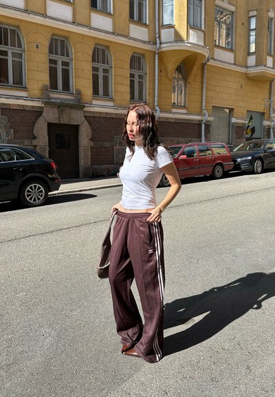 Mujer con camiseta blanca y pantalones de rayas marrones de Adidas está de pie en una calle soleada con un edificio amarillo y coches aparcados de fondo.