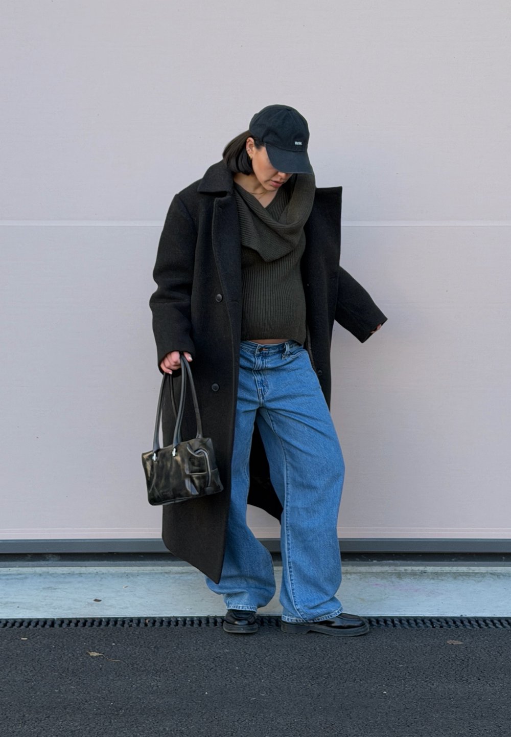 Manteau en laine noire, pull col bénitier vert foncé, jean large bleu, casquette noire, et petit sac à main noir à la texture brillante.