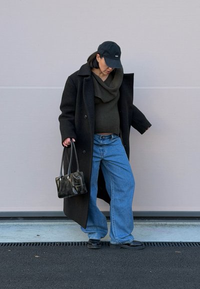 Abrigo de lana negro, suéter de cuello alto en verde oscuro, vaqueros de pierna ancha azules, gorra negra y un bolso pequeño negro con textura brillante.