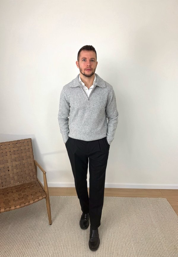 Uomo in piedi con le mani nelle tasche, indossa un maglione grigio a maglia, una camicia bianca, pantaloni neri e scarpe nere su un pavimento coperto da moquette vicino a una sedia intrecciata.