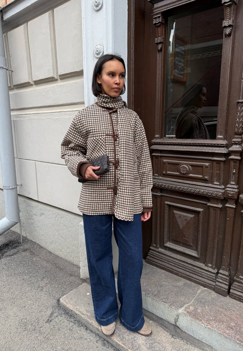 Femme debout près d'une porte en bois ornée, portant une veste à carreaux, un jean large bleu, des chaussures beiges, tenant une pochette noire tissée.