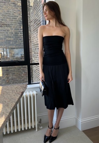 Top negro sin mangas y falda negra fluida, combinados con tacones de charol negros. Sosteniendo un pequeño bolso negro y crema.