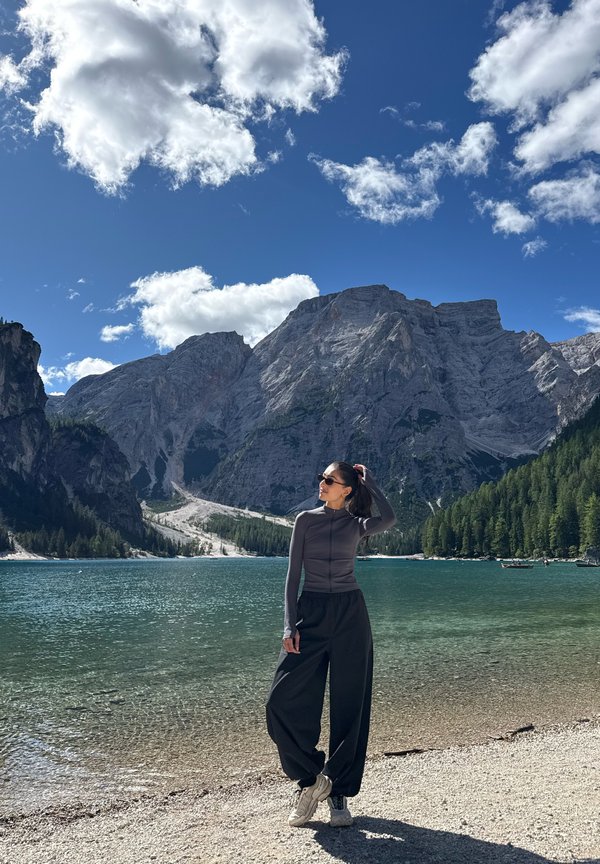 Woman wearing sunglasses and dark clothing standing by a clear lake with mountains and forest under a blue sky with scattered clouds.