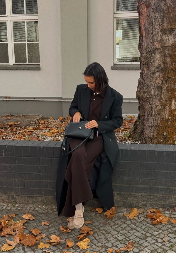 Svart frakk over en brun antrekk, sittende på en steinmur. Svart veske holdes i fanget. Området rundt har falne blader og en trestamme.