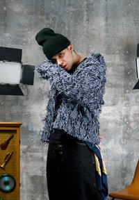Personne portant un bonnet noir et une veste à rayures bleues texturée posant devant un mur gris texturé avec des lumières de studio et un meuble en bois.