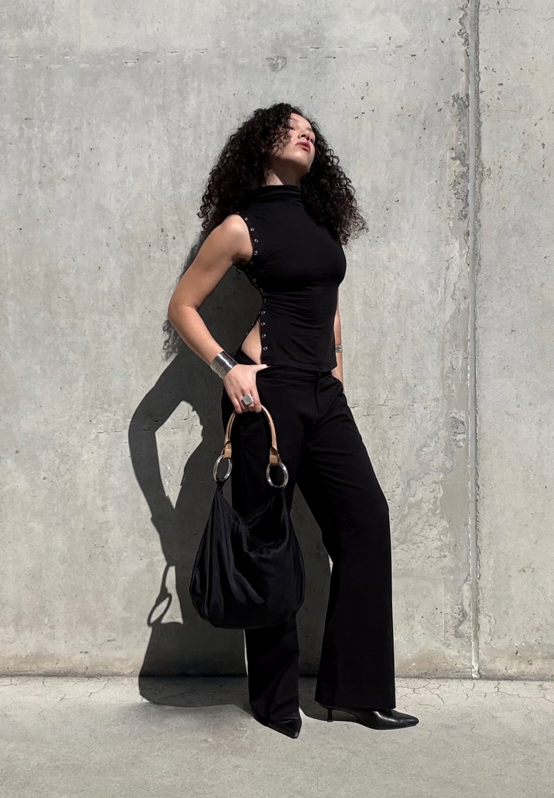 Femme aux cheveux bouclés portant un haut noir sans manches et un pantalon, tenant un sac à main noir, debout contre un mur en béton au soleil.