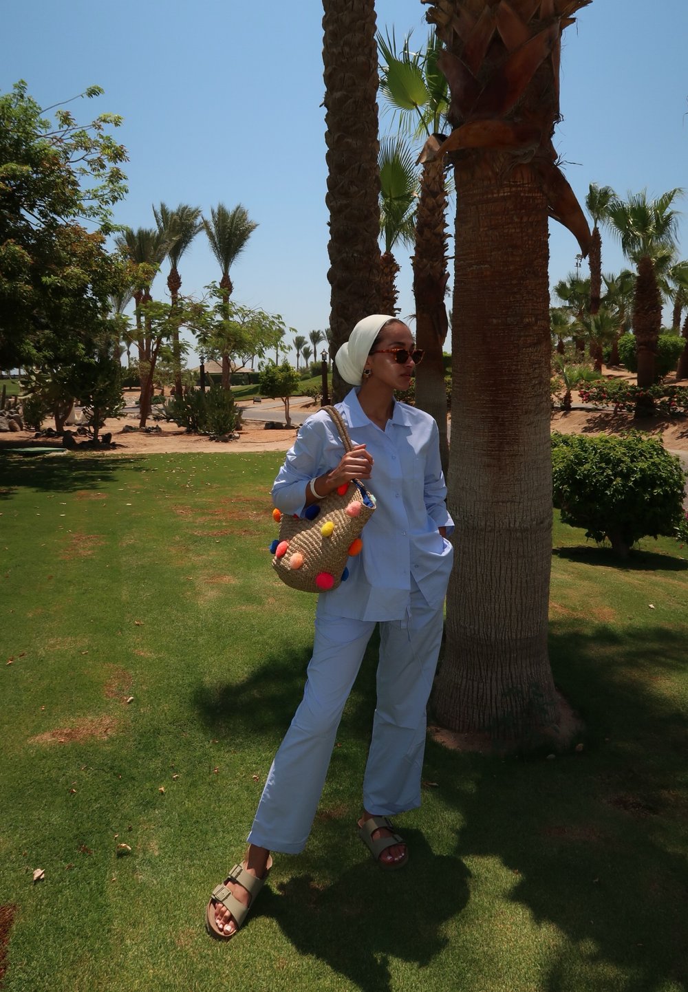 Ανοιχτό μπλε, πουκάμισο με κουμπιά και ταιριαστά παντελόνια, συνδυασμένα με μια πλεκτή τσάντα που διαθέτει πολύχρωμες πομ-πόμ. Καθισμένος δίπλα σε φοίνικες σε χορτάρι.