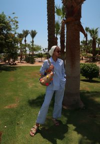 Ανοιχτό μπλε, πουκάμισο με κουμπιά και ταιριαστά παντελόνια, συνδυασμένα με μια πλεκτή τσάντα που διαθέτει πολύχρωμες πομ-πόμ. Καθισμένος δίπλα σε φοίνικες σε χορτάρι.