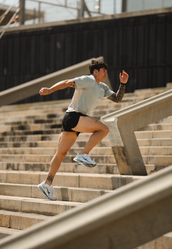Atleta con tatuaggi e maglietta azzurra che sprinta su delle scale esterne in pietra con scarpe da ginnastica durante il giorno.