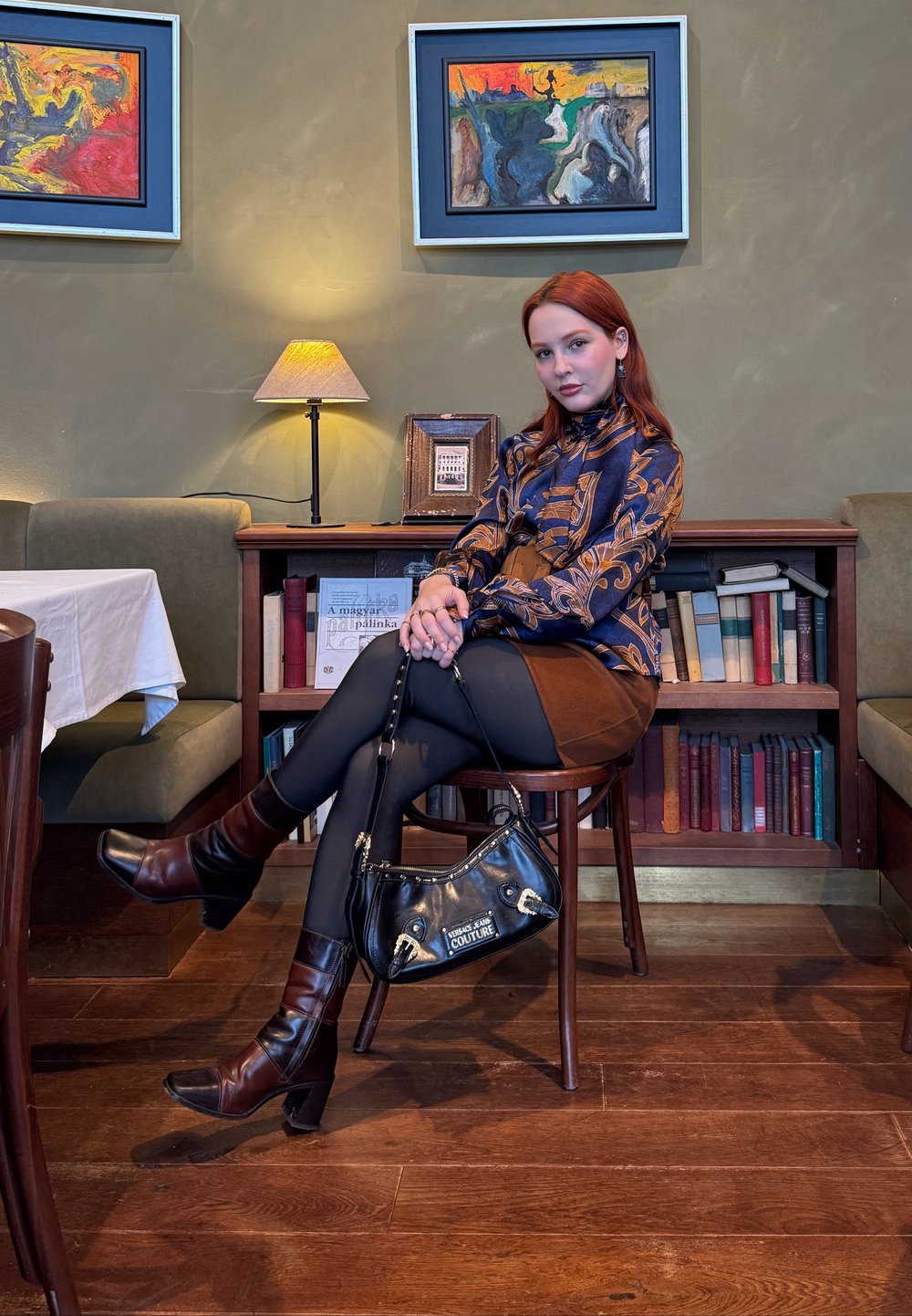 Jeune femme aux cheveux roux portant un chemisier à motifs et une jupe marron assise sur une chaise en bois, tenant un sac à main noir, dans une pièce chaleureuse avec des livres et des tableaux.