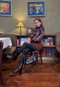 Jeune femme aux cheveux roux portant un chemisier à motifs et une jupe marron assise sur une chaise en bois, tenant un sac à main noir, dans une pièce chaleureuse avec des livres et des tableaux.