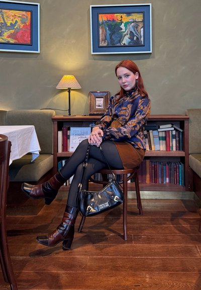 Joven con cabello rojo, blusa estampada y falda marrón, sentada en una silla de madera, sosteniendo un bolso negro, en una habitación acogedora con libros y pinturas.