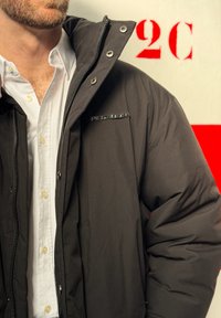 Homme portant une chemise blanche et une veste noire zippée avec boutons-pression, debout près d'un mur marqué du texte rouge « 2C ».