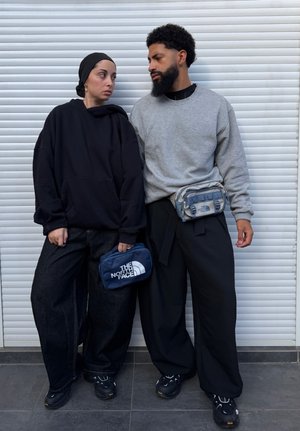 Mujer con sudadera con capucha negra, jeans anchos y pañuelo en la cabeza sosteniendo una bolsa azul, de pie junto a un hombre con sudadera gris y pantalones negros con una riñonera.