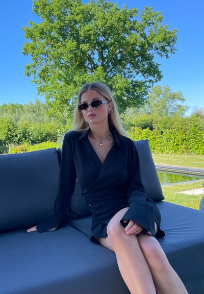 Vestido negro de envoltura con mangas largas y acampanadas, sentado en un sofá gris al aire libre. Los accesorios incluyen gafas de sol pequeñas y un collar de oro.