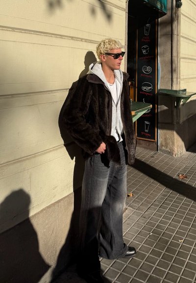 Abrigo de pelo sintético marrón oscuro superpuesto sobre una sudadera gris, combinado con jeans negros acampanados y zapatos negros. De pie contra una pared bajo la luz del sol.