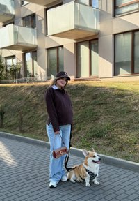 Zwangere vrouw in casual kleding en een pet staat op het trottoir en houdt een lijn vast die is bevestigd aan een zittende corgi met een harnas.
