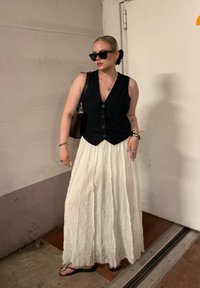 Colete preto sem mangas com detalhes em botão, saia maxi branca com efeito enrugado, sandálias pretas e óculos de sol pretos; bolsa pendurada e acessórios de joalharia.