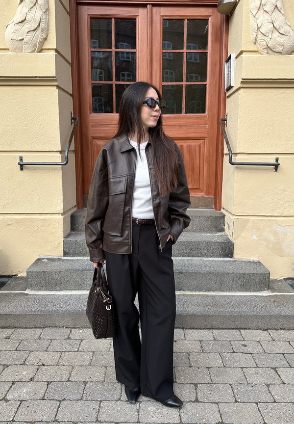 Naine pikkade tumedate juustega, kandes päikeseprille, pruuni nahktagi, valget toppi, musti laiu pükse, hoides pruuni käekotti, seisab puidust ukseastmete juures.