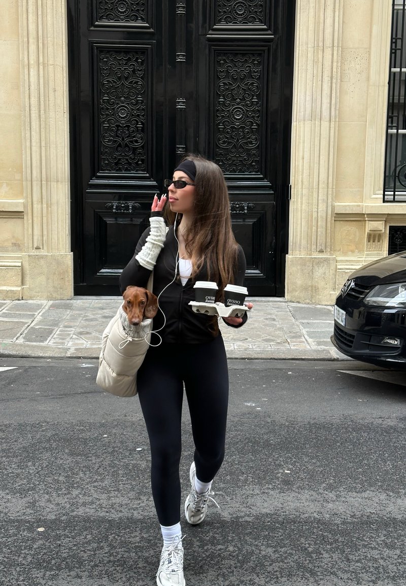 Femeie purtând haine sport negre și ochelari de soare care poartă două căni de cafea și un câine mic într-o geantă bej purtată pe umăr pe o stradă din oraș.