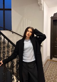 Schwarzer langer Mantel über einem hellblauen Oberteil, kombiniert mit einer dunklen Hose. Im Hintergrund ein kunstvolles schwarzes Geländer und ein Bodenfliesenmuster.