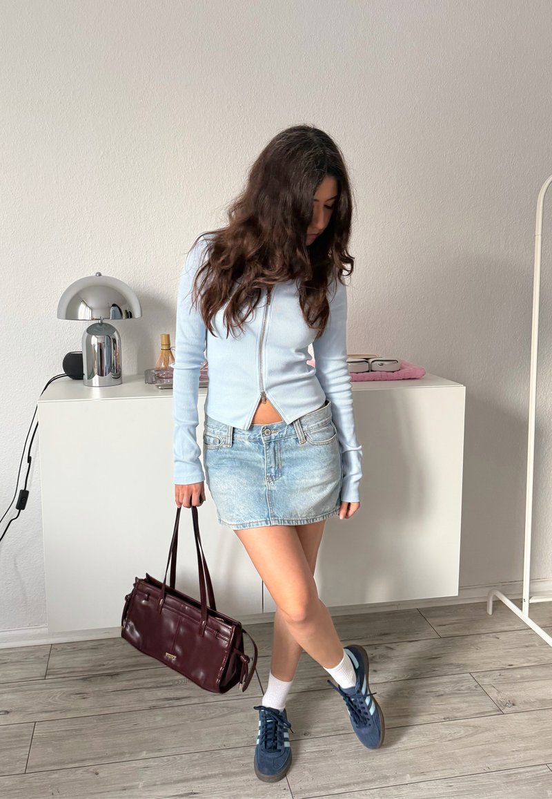 Žena s dlhými vlasmi, oblečená v svetlomodrom top s na zips, rifľovej mini sukni, bielych ponožkách a modrých teniskách, držiaca tmavohnedú kabelku v interiéri.