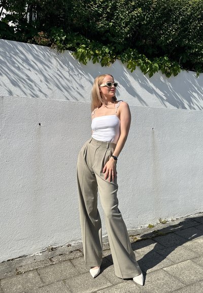 Top sin mangas ajustado blanco con tirantes finos, combinado con pantalones de tiro alto en color beige claro. El calzado son tacones blancos puntiagudos. El fondo es una pared blanca.