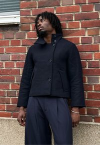 Granatowa kurtka z mieszanki wełny, z wysokim kołnierzem i zapięciem na guziki z przodu, wyposażona w dwie boczne kieszenie i krótki fason, na tle ceglanej ściany.