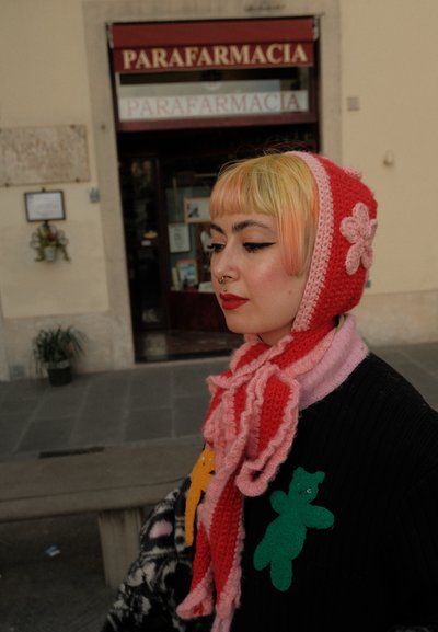 Persona con cabello multicolor que lleva un pañuelo de cabeza de punto rojo y rosa y un suéter negro con parches de osos verdes y naranjas, de pie al aire libre.