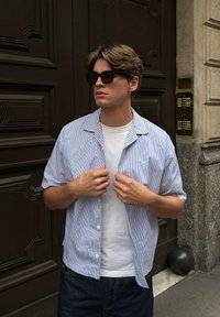 Camicia a maniche corte con bottoni, a strisce blu e bianche, indossata sopra una maglietta bianca. Occhiali da sole scuri, in piedi davanti a una porta in legno texturizzata.