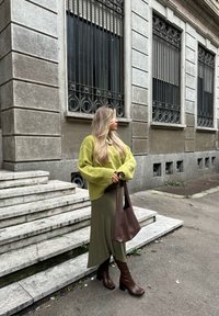 Maglione verde oversize, gonna oliva, stivali marroni fino al ginocchio e borsa a tracolla marrone. Presenta un motivo a maglia testurizzata e un look a strati. Scale sullo sfondo.