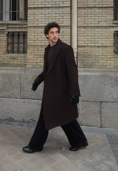 Abrigo largo de color marrón oscuro sobre un atuendo negro, con guantes y zapatos negros. La figura camina por una calle adoquinada cerca de un muro de ladrillo.