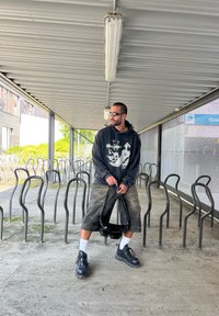 Nero felpa con stampa grafica, pantaloni corti grigi larghi, scarpe nere, calzini bianchi e una borsa nera; in piedi tra sedie di metallo in un corridoio.