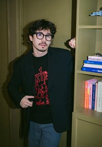 Young man with curly hair and glasses wears a black blazer and graphic t-shirt, standing next to a bookshelf with colourful books.