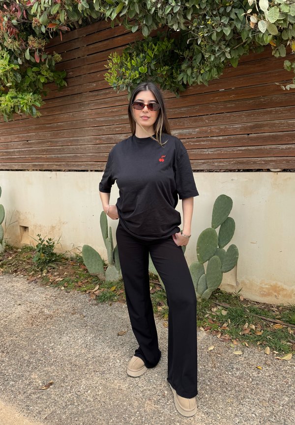 Junge Frau mit Sonnenbrille, schwarzem T-Shirt mit Kirsch-Logo, schwarzer Hose und beigen Schuhen steht draußen neben einem Holzzaun und Kakteen.
