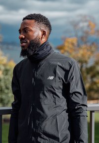 Bearded man wearing a black New Balance jacket and neck gaiter standing outdoors with blurred autumn trees in the background.