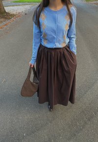 Lichtblauwe cardigan met ruitpatroon, bruin plissérok, zwarte hakken en een bruine geweven handtas. Buitenomgeving op het trottoir.