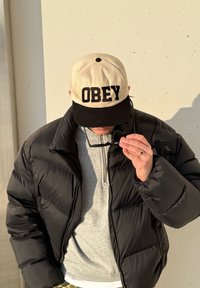 Personne portant une doudoune noire, un pull gris à zip, une casquette noire et beige avec l'inscription "OBEY", tenant des lunettes de soleil près de son visage, tête inclinée vers le bas.