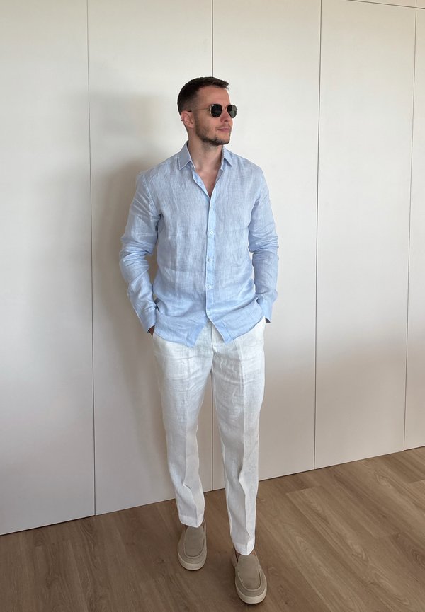 Hombre con camisa azul claro de manga larga, pantalones blancos, zapatos beige y gafas de sol, de pie en el interior junto a una pared lisa y suelo de madera.