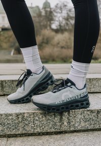 Grijze hardloopschoenen met een ademend mesh bovenwerk, zwarte accenten, Cloudtec-logo en een dikke, gedempte zool op een gestructureerd oppervlak.