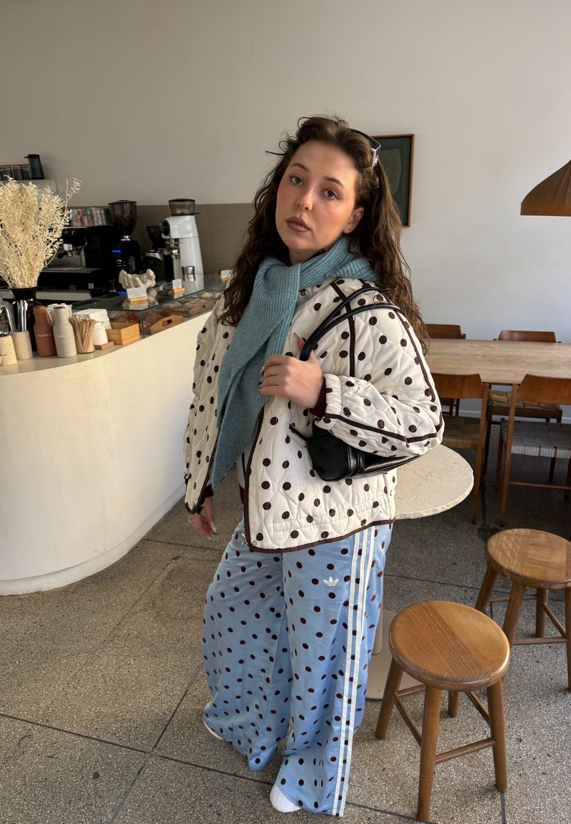 Jeune femme portant une veste à pois et un pantalon avec une écharpe bleue se tient dans un café près de tabourets en bois et d'une table ronde.