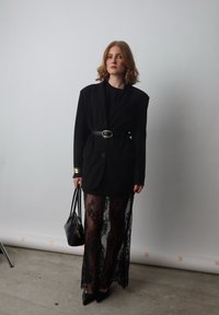 Nero blazer oversize con dettagli dorati, gonna trasparente in pizzo nero, tacchi a stiletto e borsa nera, con intricati motivi in pizzo.