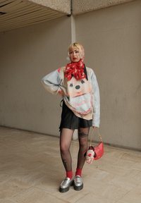 Strickpullover mit einem lächelnden Gesichtsmotiv in Pastellfarben, kombiniert mit einem schwarzen Rock, roten Socken, silbernen Plateauschuhen und einer roten Handtasche.