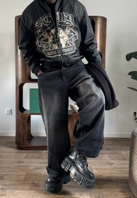 Personne portant une veste noire à motifs, un jean noir à jambes larges et des bottes noires épaisses, debout à l'intérieur sur un sol en bois près des étagères.