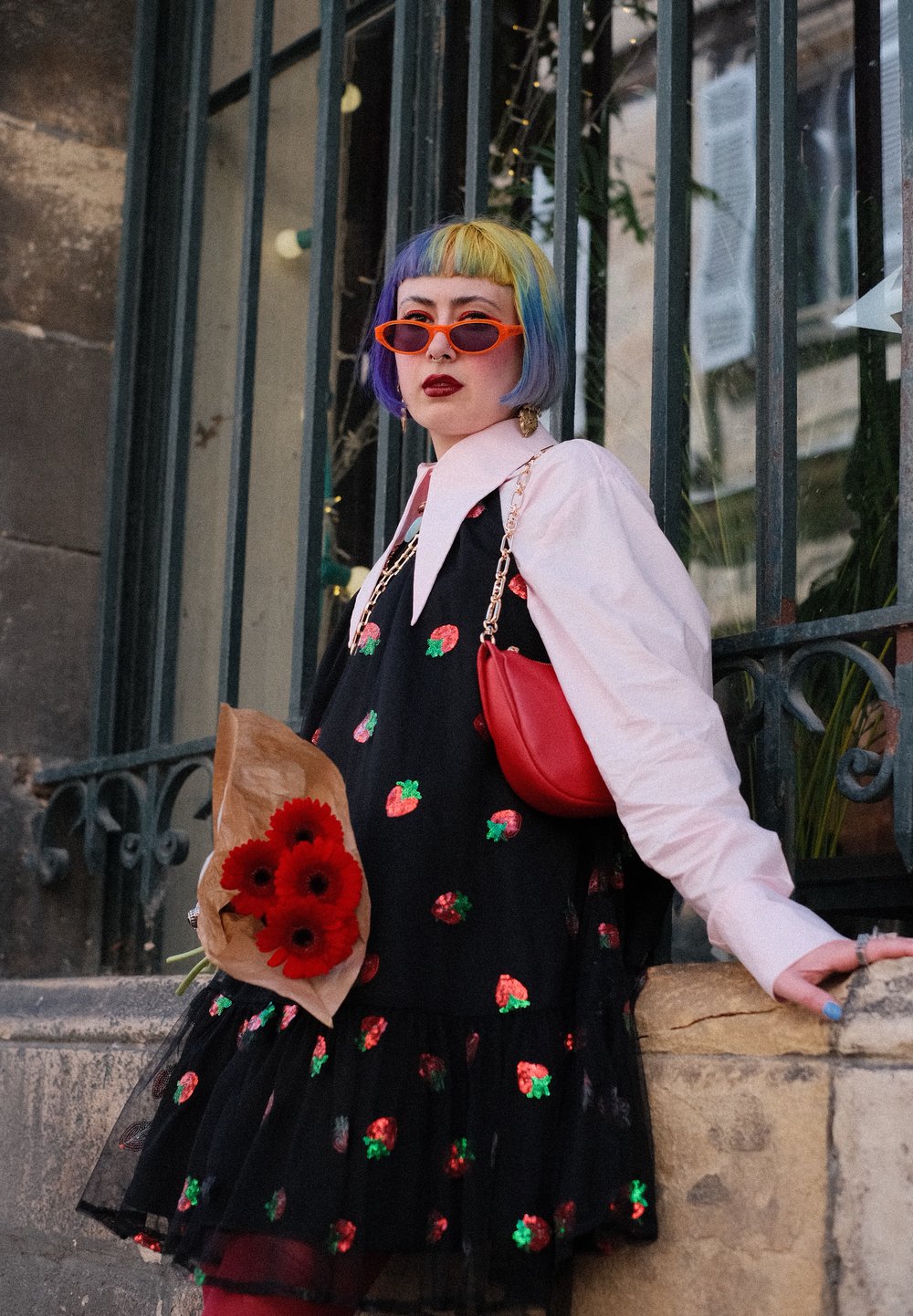 Robe noire avec des motifs de fraises, col blanc, sac à épaule rouge, coupe de cheveux bob multicolore, et un bouquet de fleurs rouges.