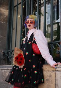 Robe noire avec des motifs de fraises, col blanc, sac à épaule rouge, coupe de cheveux bob multicolore, et un bouquet de fleurs rouges.